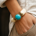 Bracciale rigido in osso bianco con grande turchese e dettagli in ottone, indossato al polso.